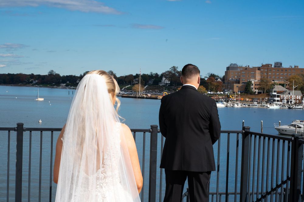 A wedding couple stands at a waterfront railing overlooking a marina and harbor on a sunny day.