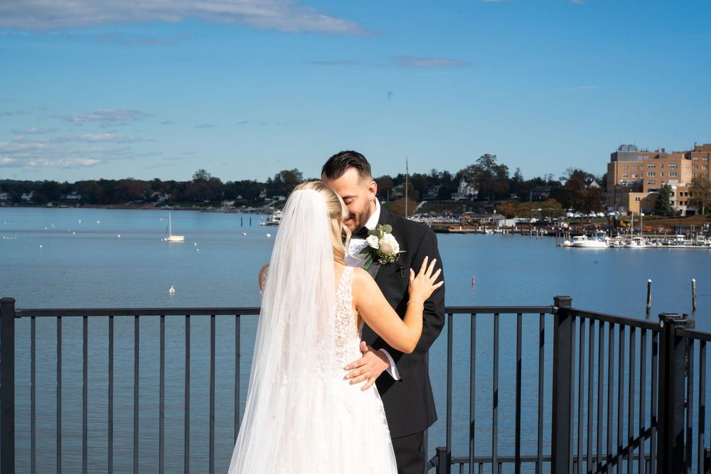 Couple embracing on a waterfront dock with boats and buildings visible in the background on a sunny day.