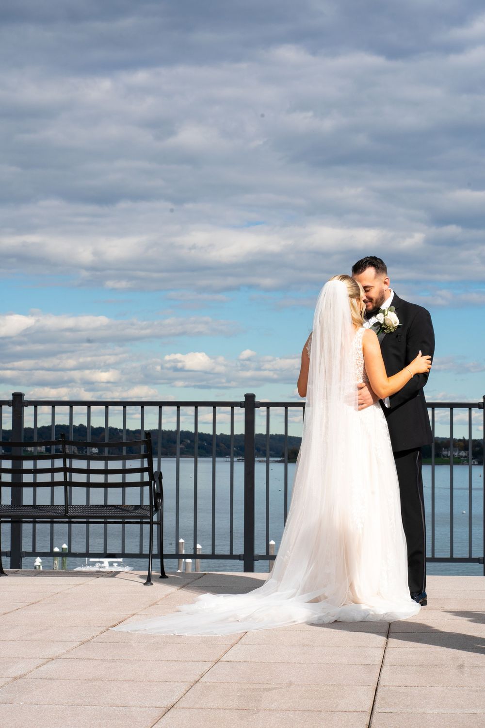A couple shares an intimate moment on a waterfront terrace with a cloudy sky and railing in the background.