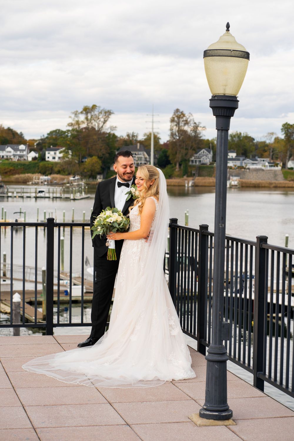 A romantic wedding couple shares an intimate moment on a waterfront deck with boats and houses in the background.