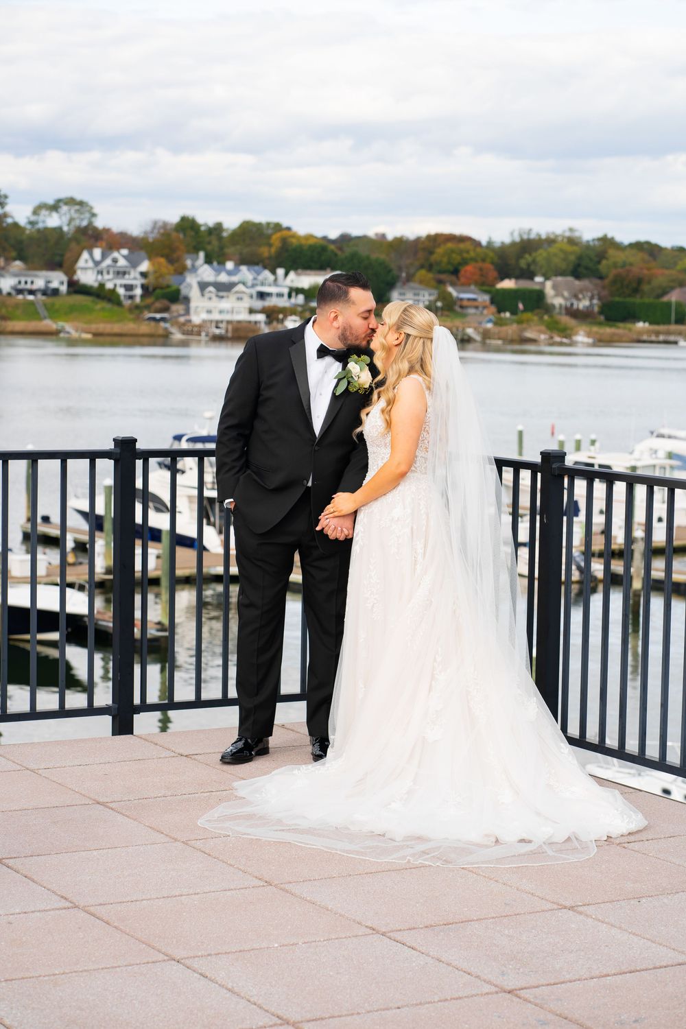 A wedding couple embraces on a marina boardwalk overlooking a scenic harbor with autumn trees in the distance.