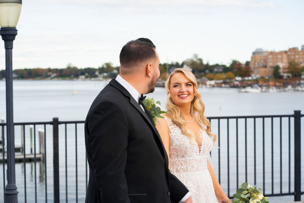 A wedding couple and their bridal party stand together on a waterfront pier during a sunny outdoor celebration.