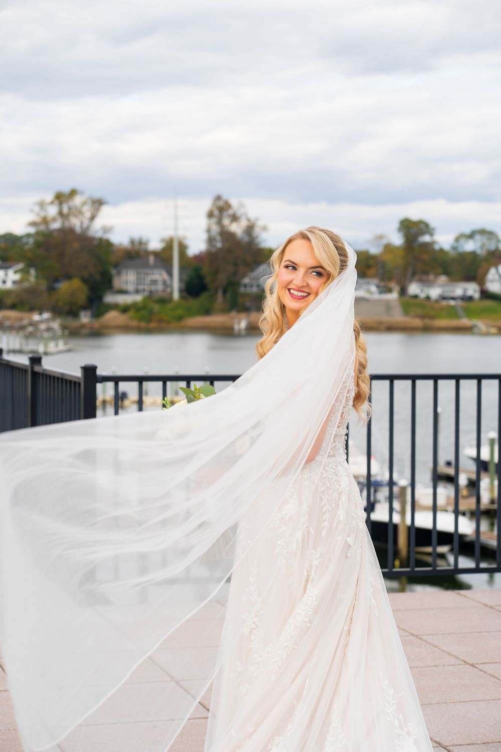 A bride in a flowing white wedding dress and veil stands on a waterfront deck with boats and trees in the background.