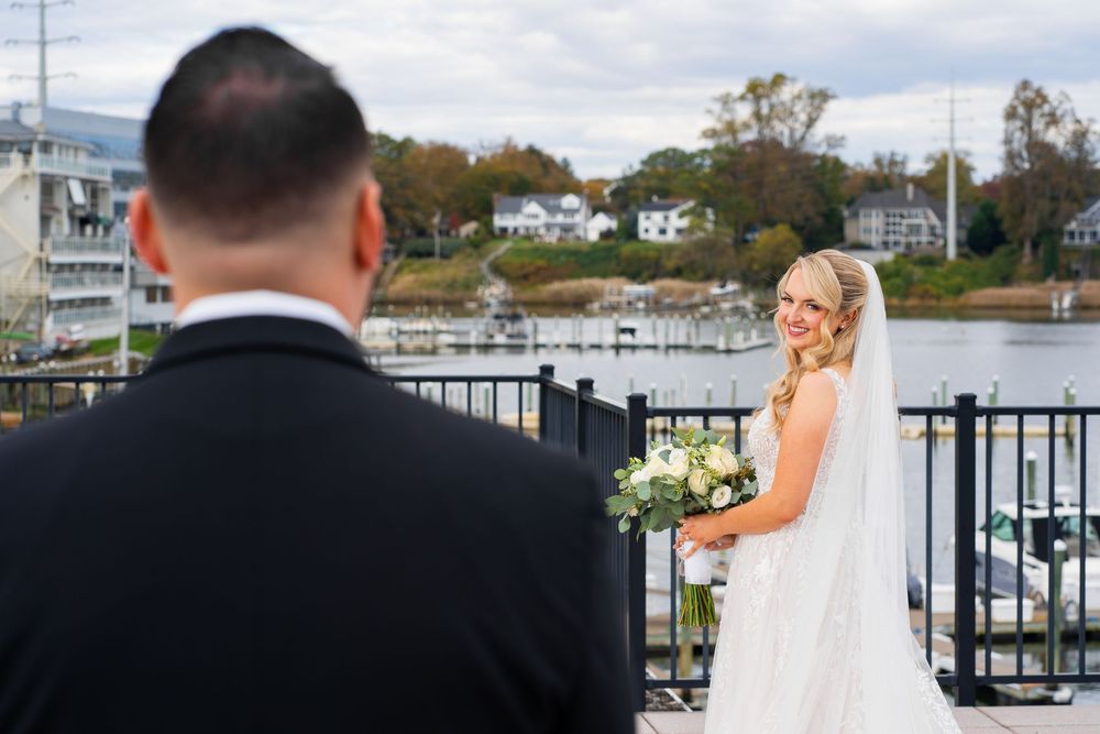 A couple in formal wedding attire stands at a waterfront dock overlooking a marina on their special day.