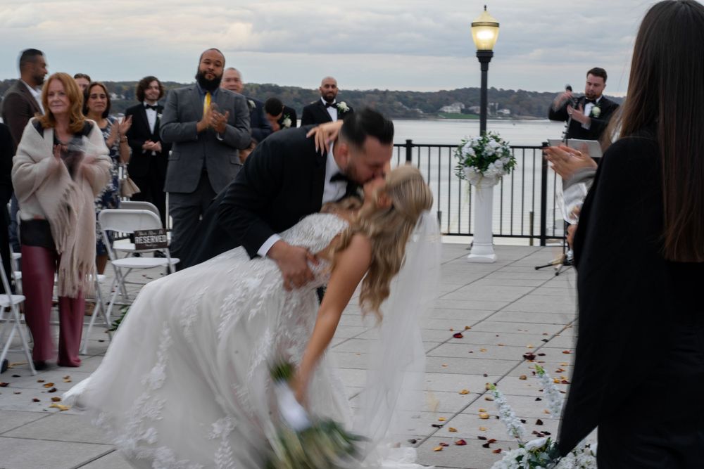 A couple shares a romantic dip kiss during their waterfront wedding ceremony as guests look on.