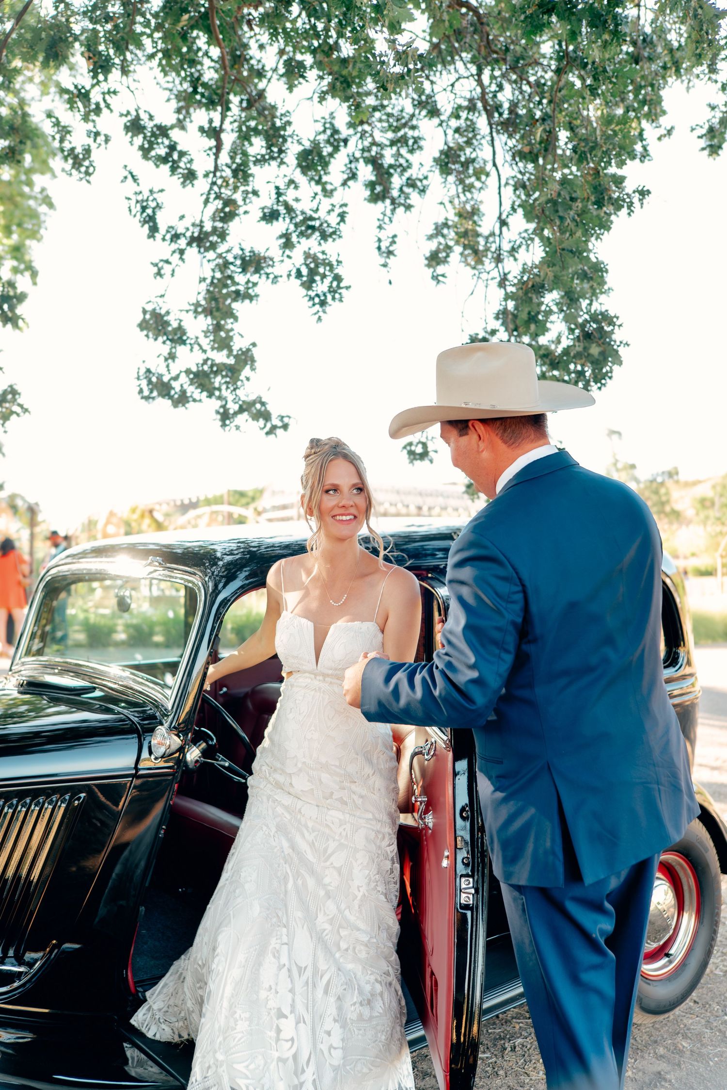 Wedding guests share joyful laughter during an outdoor reception under trees on a sunny day.