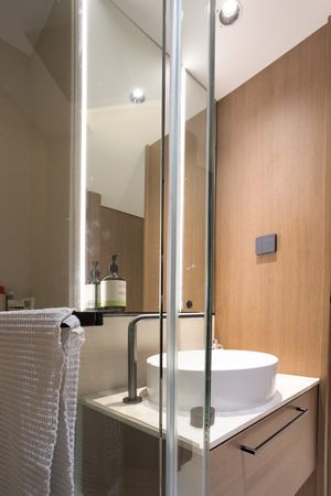 Compact bathroom with round white vessel sink, illuminated mirror cabinet, wood accent wall, and glass partition.