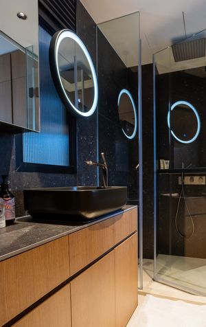 Luxury bathroom with black vessel sink, wood vanity, backlit oval mirrors, dark marble walls, and glass shower.