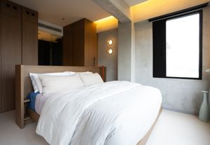 Serene minimalist bedroom with white duvet, concrete walls, warm wood wardrobe, and soft ambient ceiling lighting.