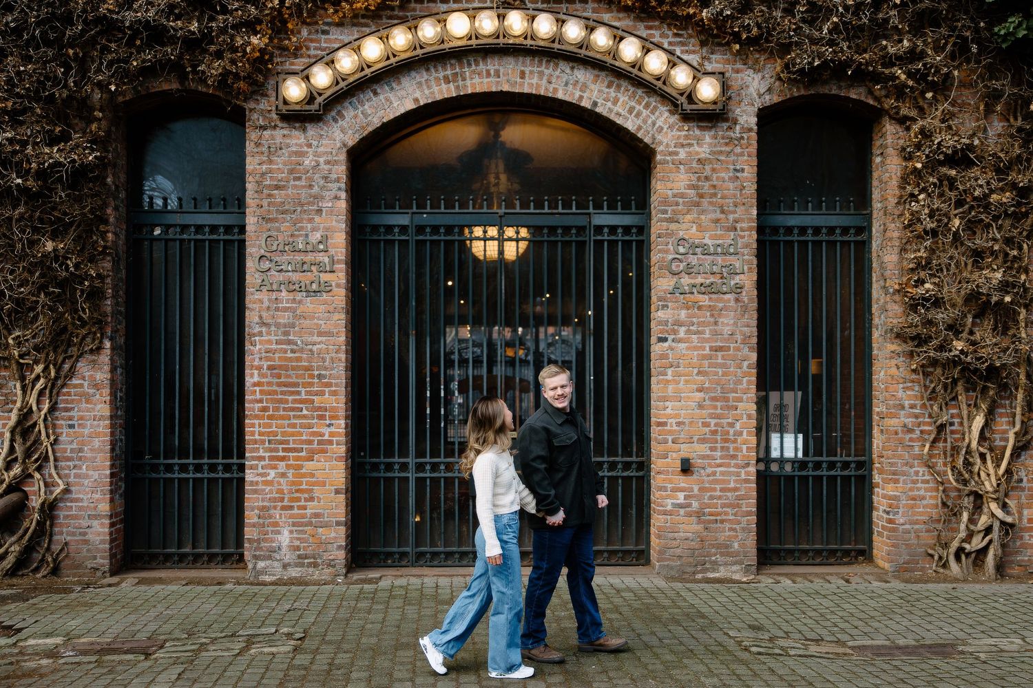 A couple walks past an arched brick entrance with iron gates and warm lighting at dusk.