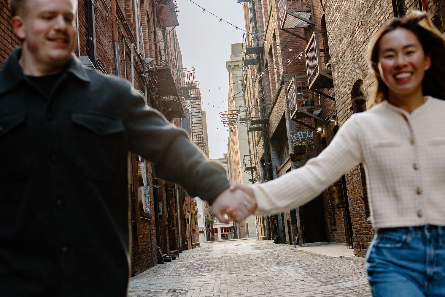 A group of friends holds hands while walking down a brick alley with string lights overhead in an urban setting.