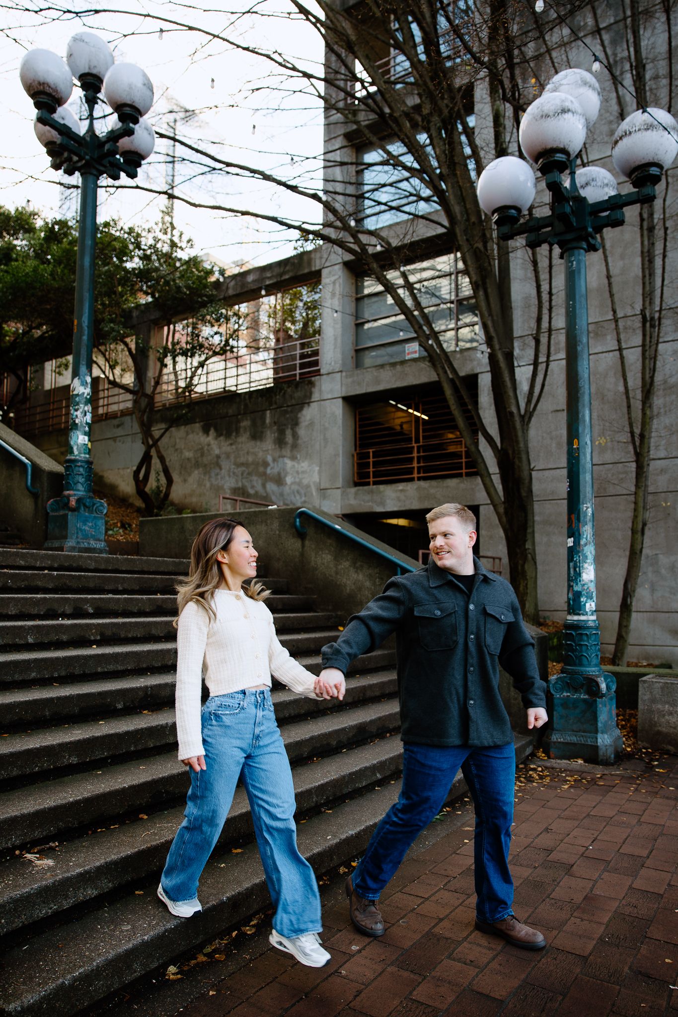 A couple in casual outfits playfully interacts on brick steps with vintage-style lamp posts in an urban setting.