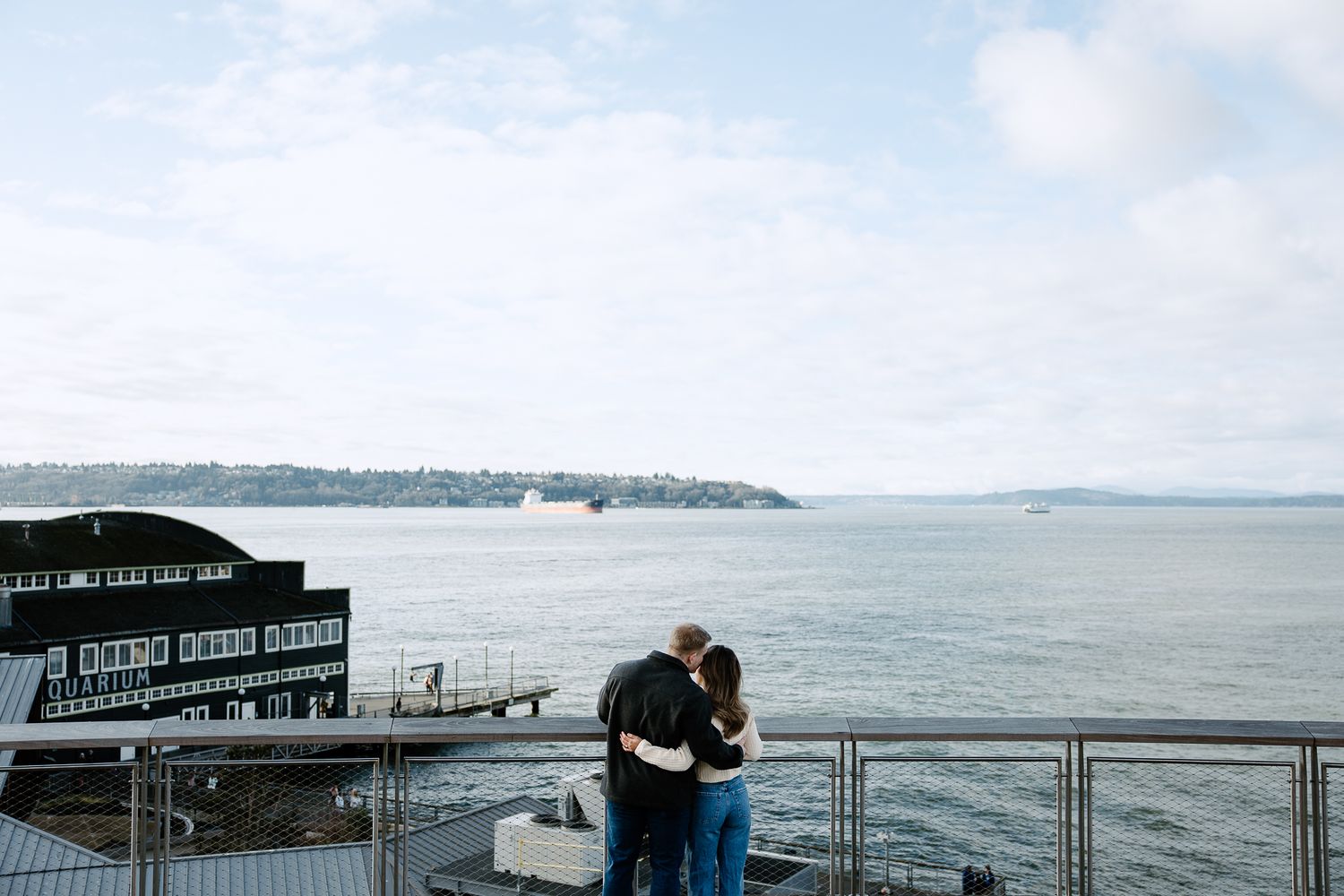 A couple embraces on a waterfront deck overlooking a scenic bay on an overcast day.