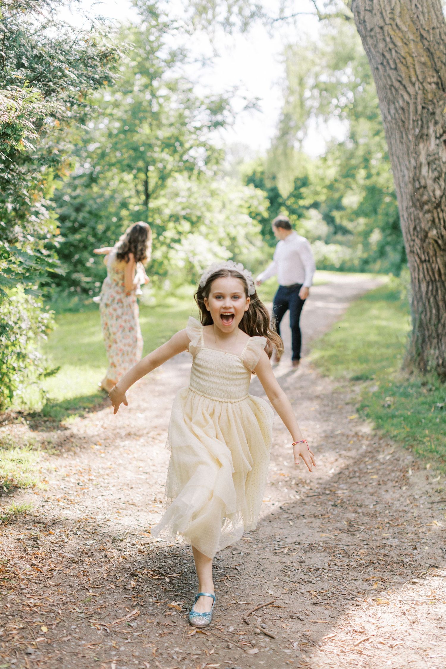 A child in a cream-colored dress runs joyfully down a sunlit garden path while others walk in the background.