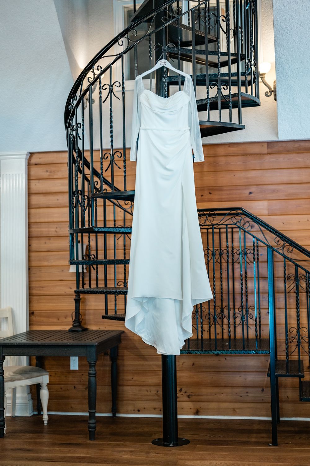 White wedding dress hangs elegantly on display by a spiral staircase with wrought iron railings.