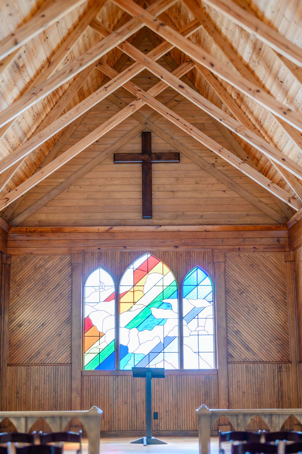 Wooden chapel interior with exposed beams and colorful stained glass windows beneath cross.