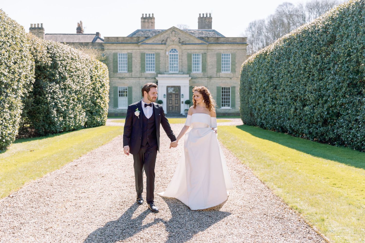 Elegant couple walking along a gravel path beside a grand historic mansion with manicured hedges and gardens.