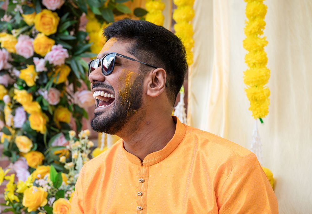 Person in orange traditional attire smiles joyfully against a backdrop of yellow and pink floral decorations.