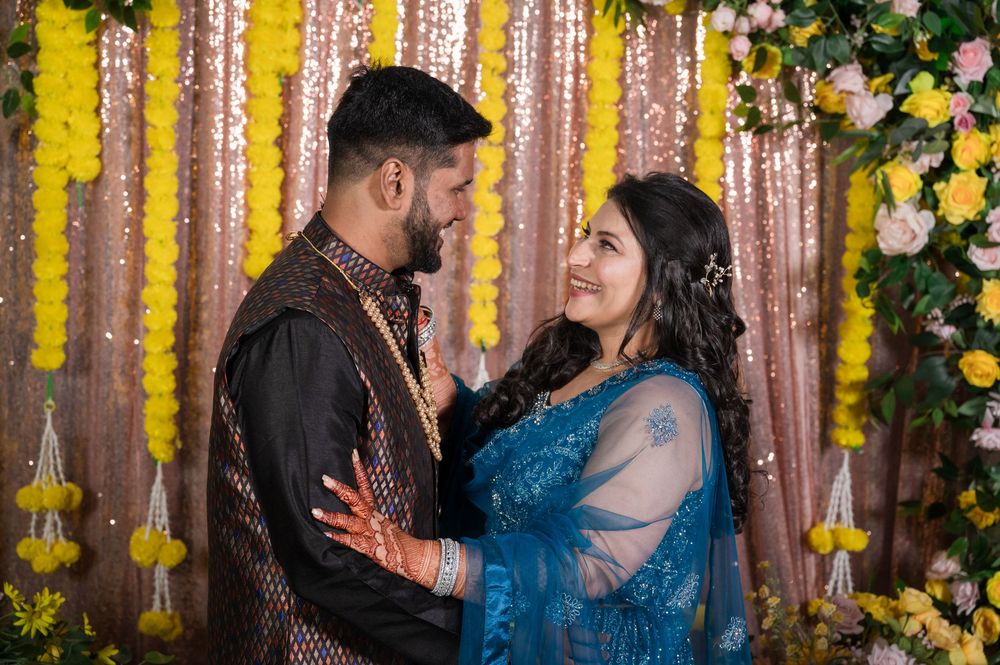 A couple in traditional Indian attire embrace at a wedding celebration with floral decorations and a rose gold sequin backdrop.