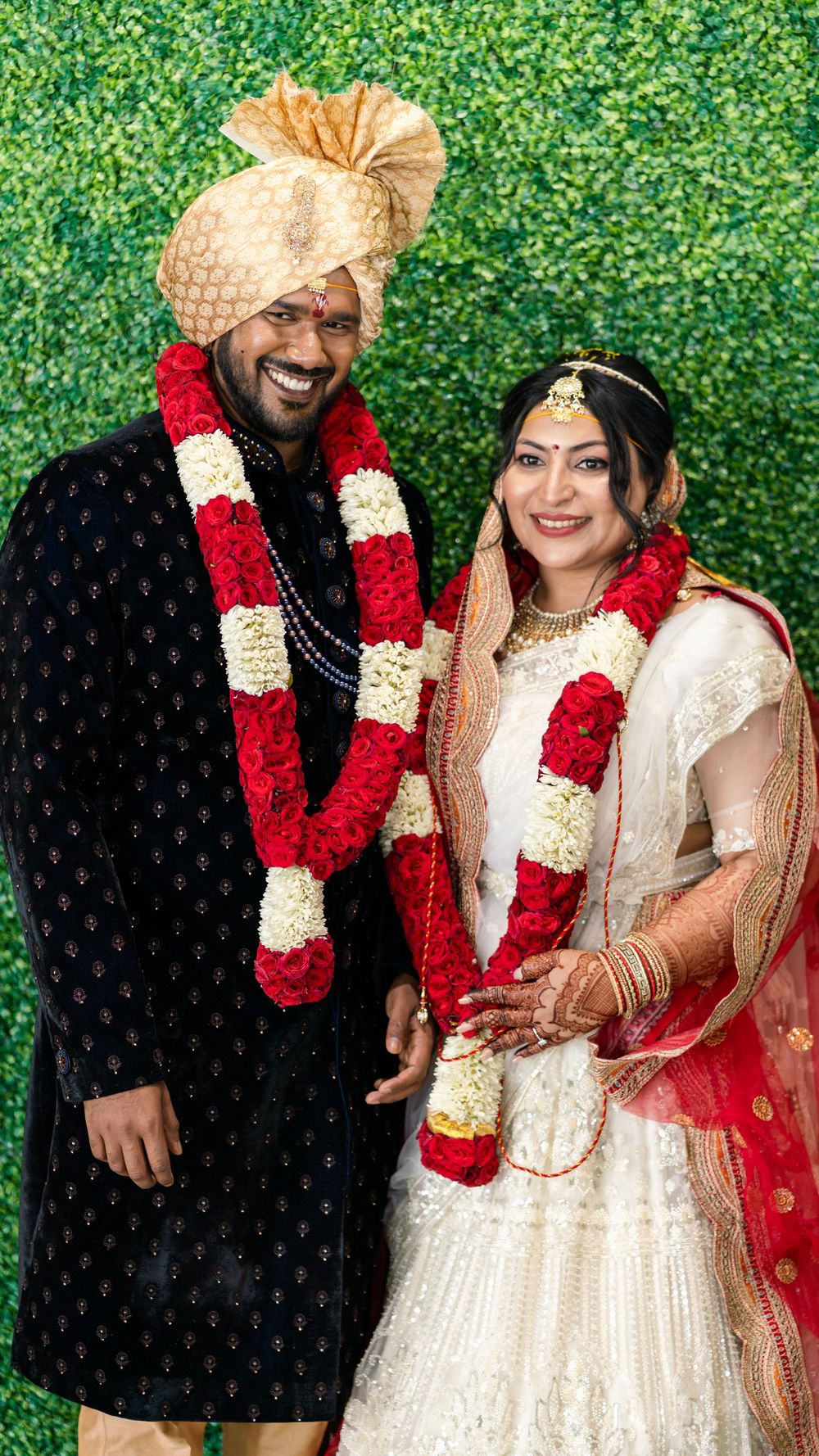 A couple in traditional Indian wedding attire with red garlands pose together against a green hedge backdrop.