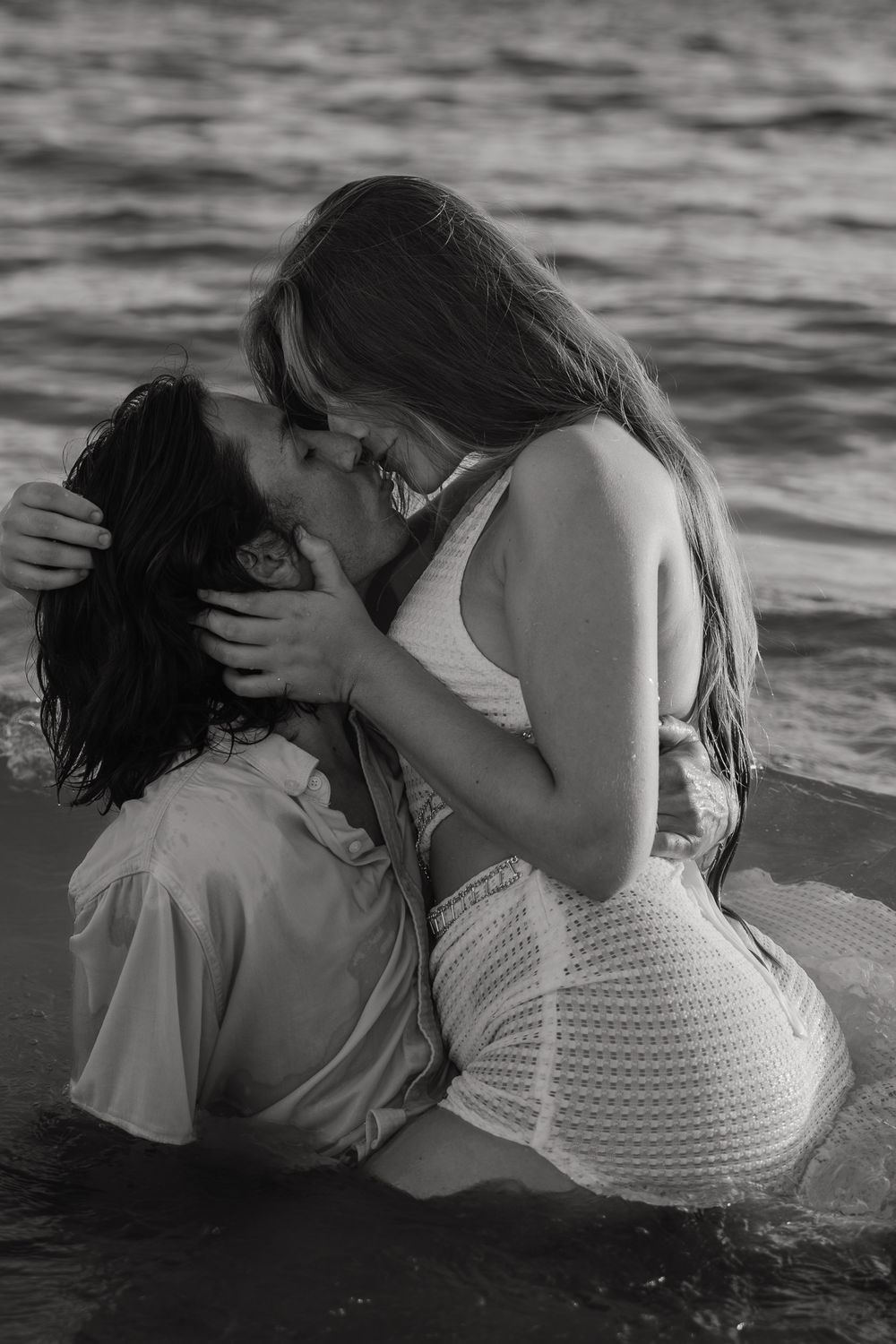 A romantic black and white photo of a couple embracing and kissing in shallow ocean water at sunset in upham beach, florida.