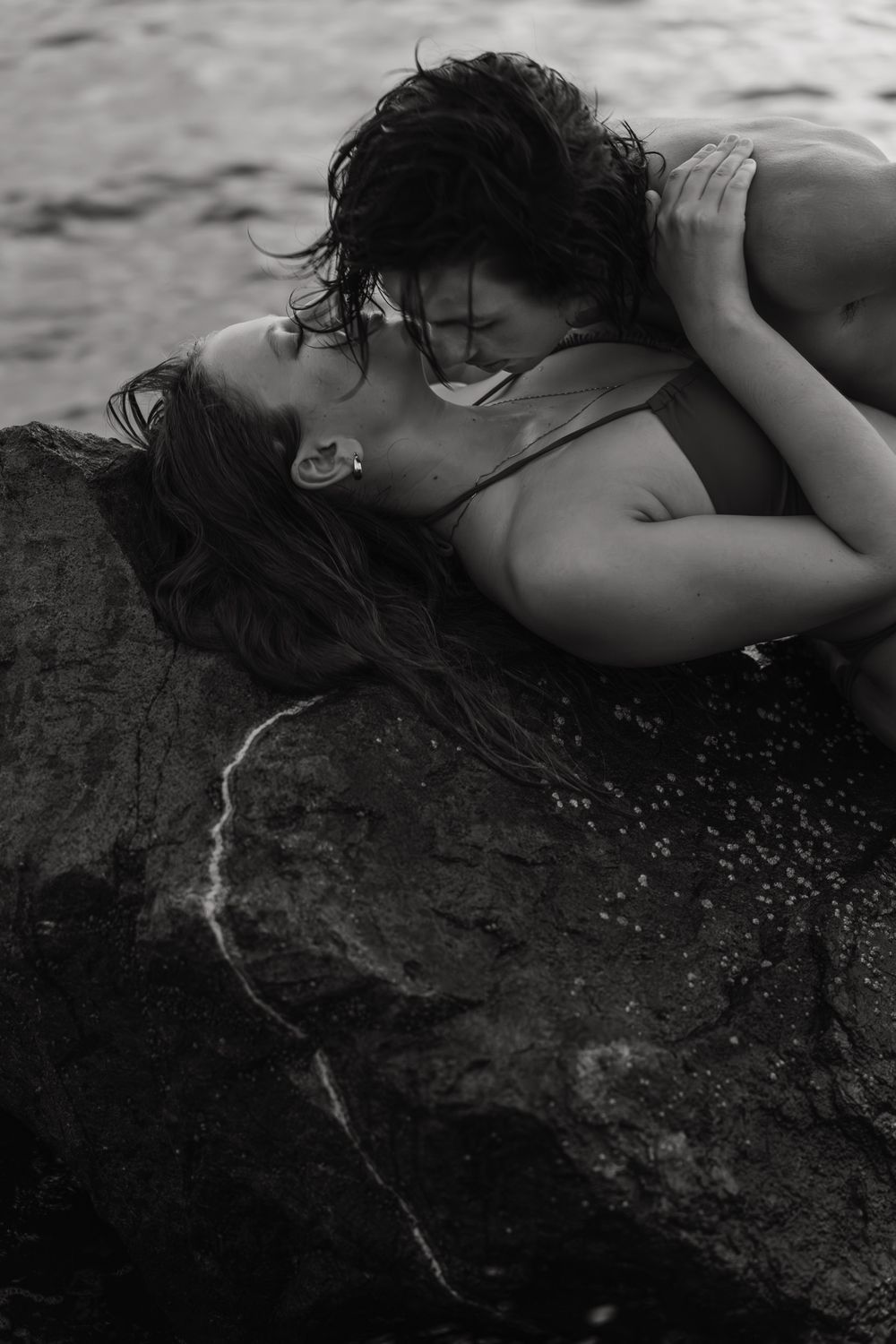 A black and white intimate photograph of a couple embracing on wet rocks near water at upham beach, florida