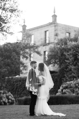 A romantic black and white wedding photo of newlyweds standing on a lawn in front of a stately building.