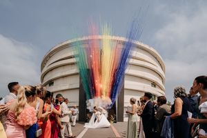 A panoramic sequence of wedding guests tossing colorful smoke bombs outside a domed building at sunset.