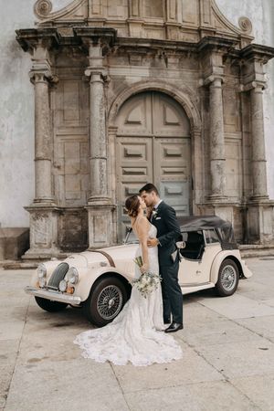 A couple poses with a classic vintage car against a historic stone archway in an elegant wedding photoshoot.