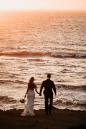 Silhouettes of newlyweds walking along beach at sunset with orange sky and waves.
