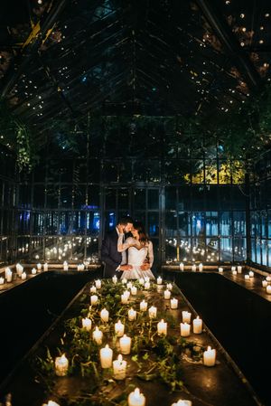 A romantic candlelit greenhouse wedding scene with glowing candles lining the aisle and twinkling lights overhead.
