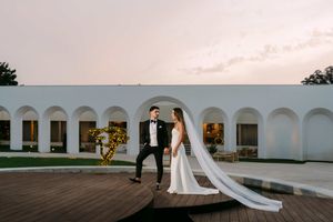 Elegant wedding couple poses against white arched colonnade at sunset.