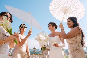 Wedding party holds white parasols against bright blue sky at beach celebration.