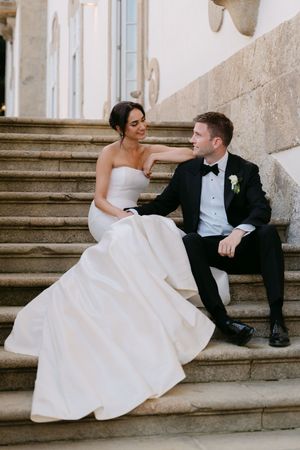 Newlyweds in formal attire relaxing on grand stone staircase at historic venue.