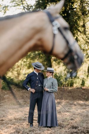 A couple in vintage early 1900s attire with straw hats stand beside a brown horse in a pastoral setting.