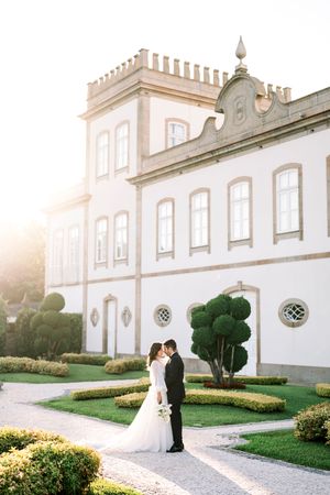 Elegant wedding couple poses outside grand white mansion with manicured gardens at sunset.