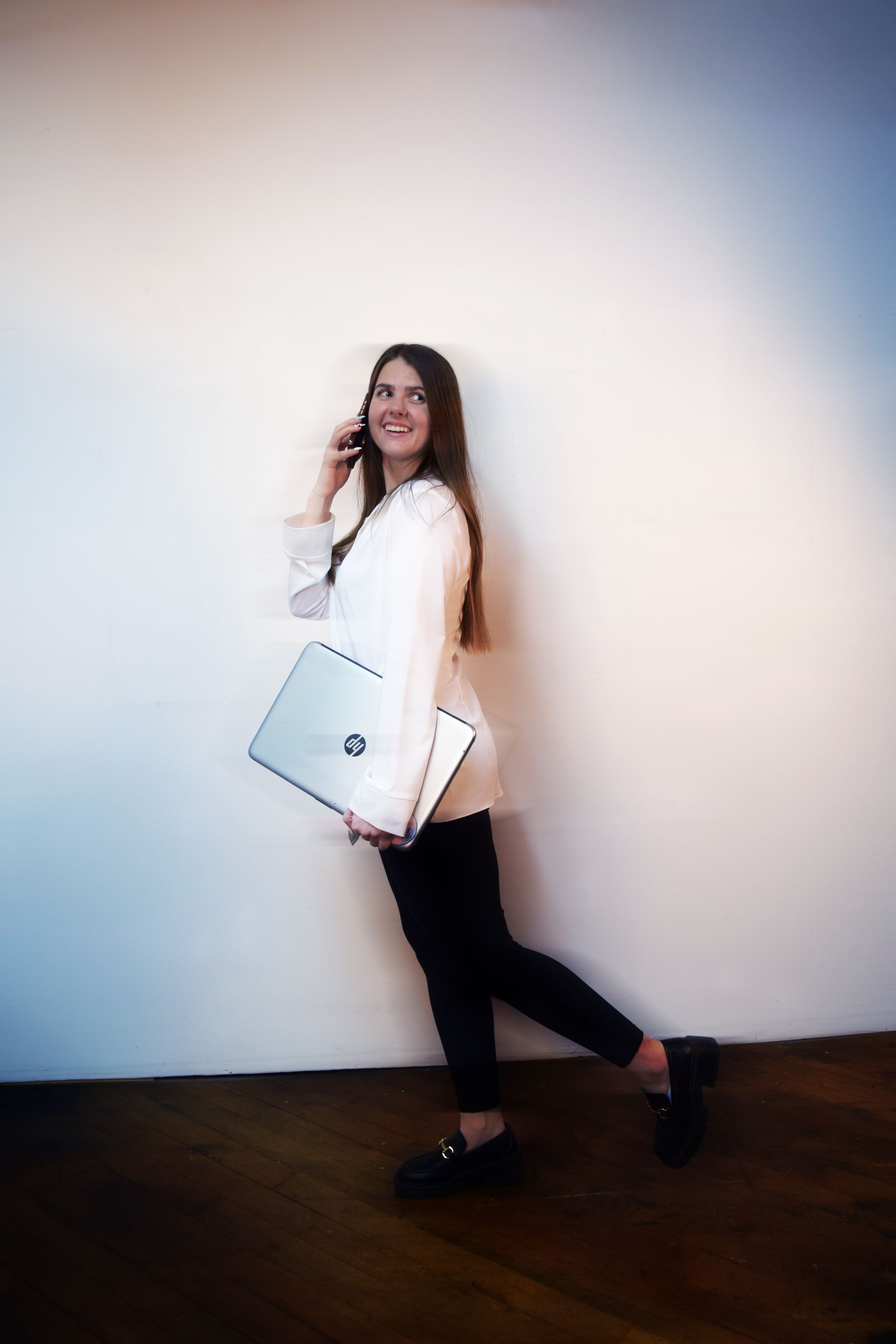 A person in black pants and white top poses playfully with a white laptop against a light-colored wall.