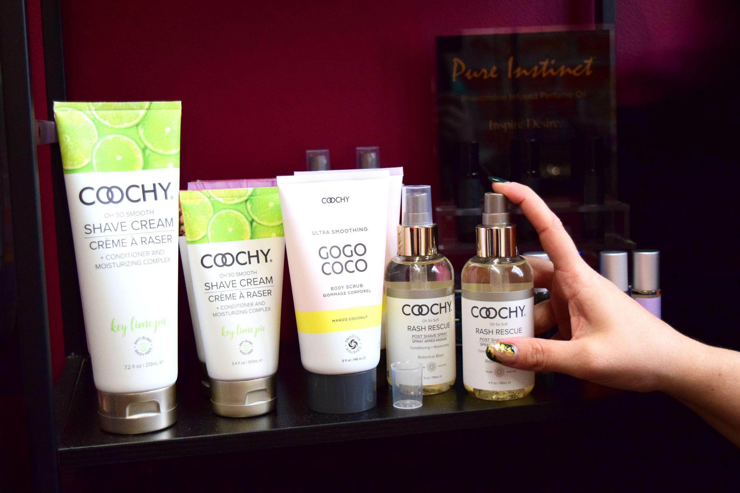 A hand reaches for natural beauty products from the Coochy brand displayed on a dark shelf with burgundy background.