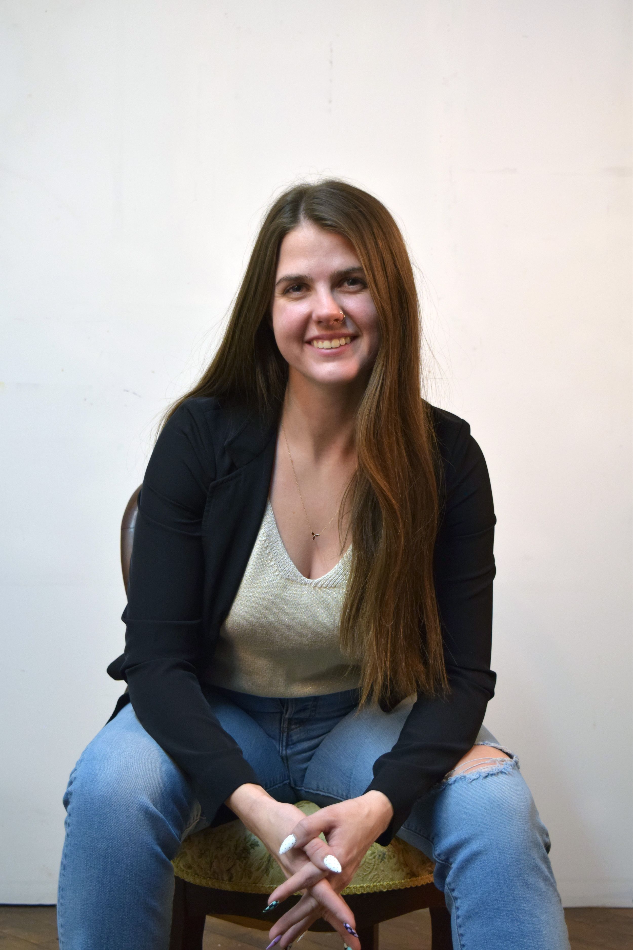 A person in a black blazer and jeans sits casually on a chair against a white wall.