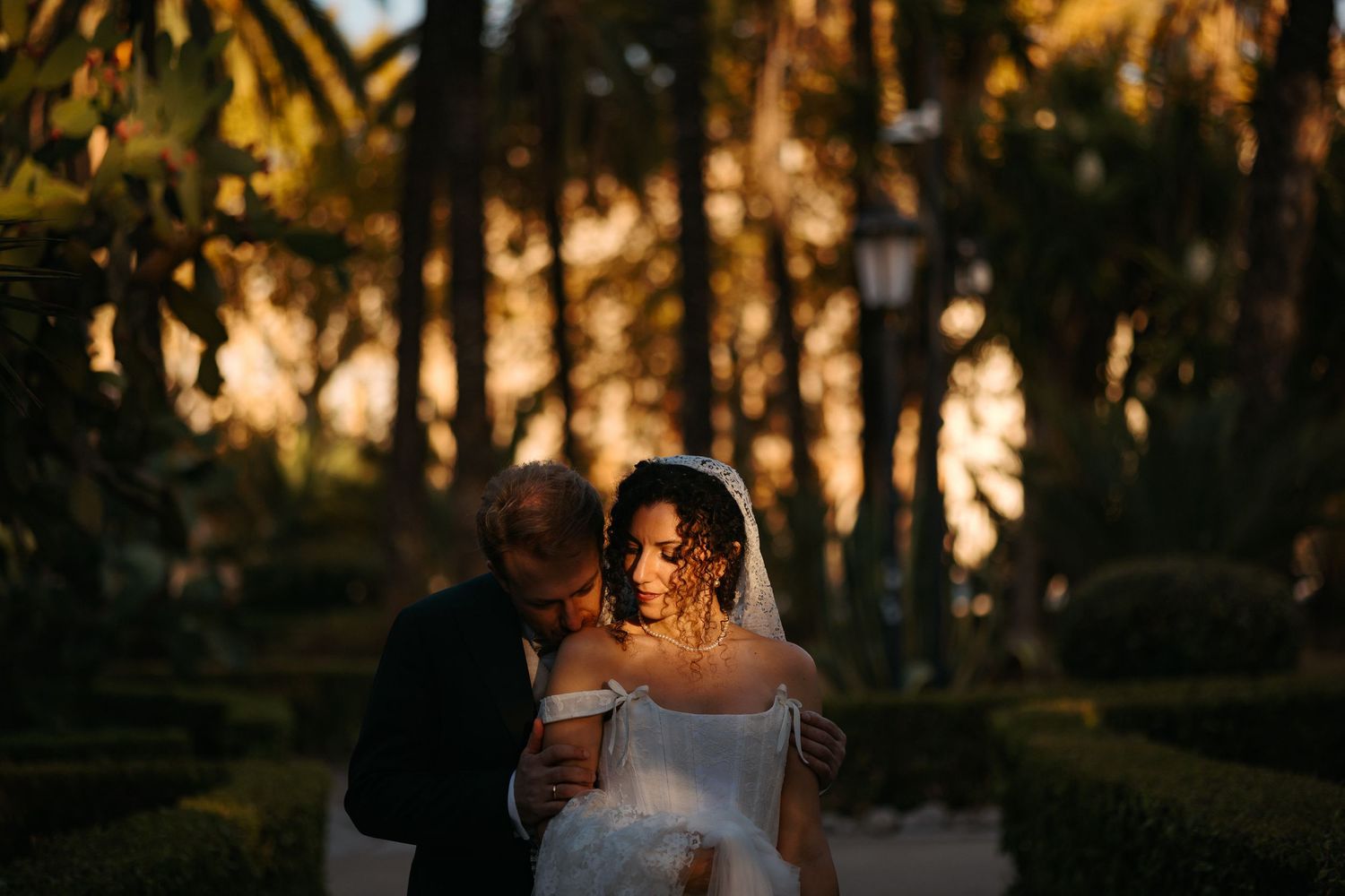 A couple shares an intimate moment at sunset in a garden with palm trees silhouetted in the golden light.