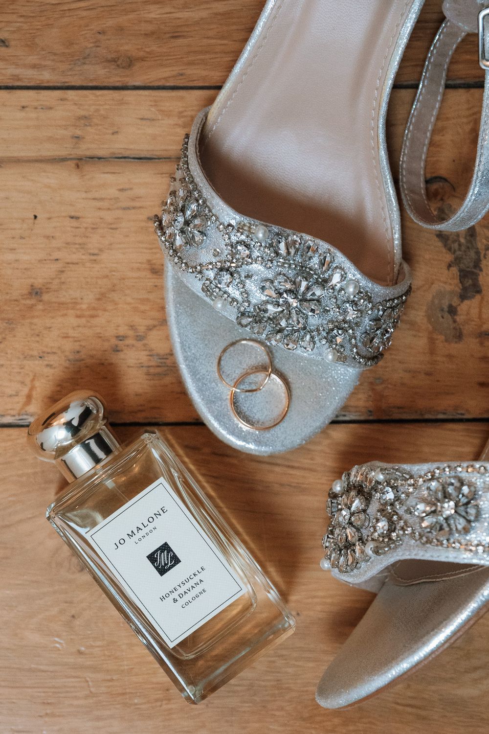 Elegant silver bridal shoe with crystal embellishments displayed with wedding rings and Jo Malone perfume on wooden floor.