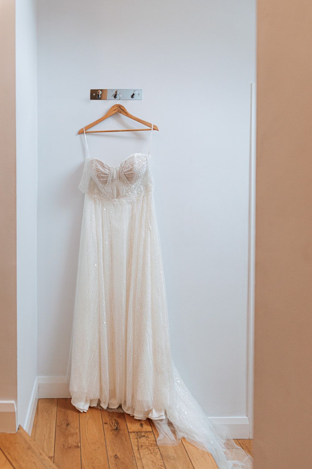 A white strapless wedding dress with lace details hangs on a wooden hanger against a white wall with hardwood flooring.