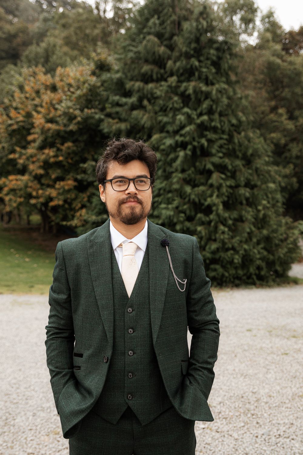 A person in a dark green three-piece suit and glasses poses outdoors against an autumn forest background.