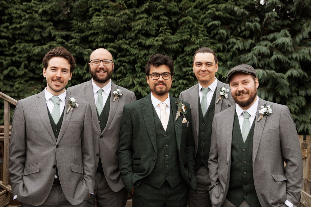 A wedding party of five groomsmen wearing coordinated gray and dark green suits standing together outdoors.