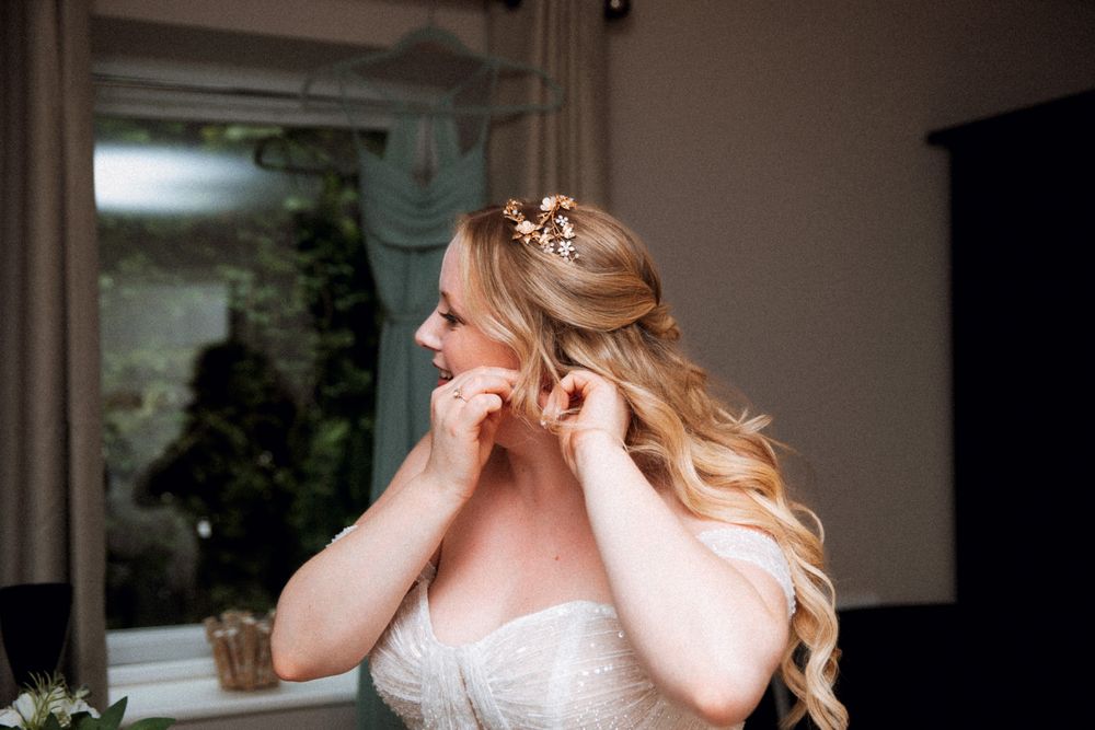A bride in a white lace gown puts on earrings while getting ready, with long wavy blonde hair adorned with delicate flowers.
