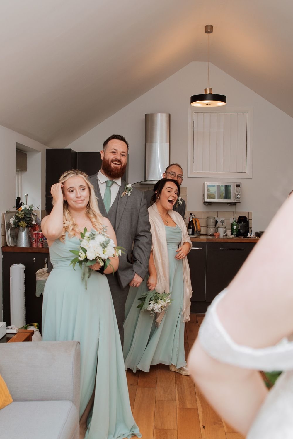 Wedding party members in sage green attire share a candid laughing moment together in a modern kitchen setting.