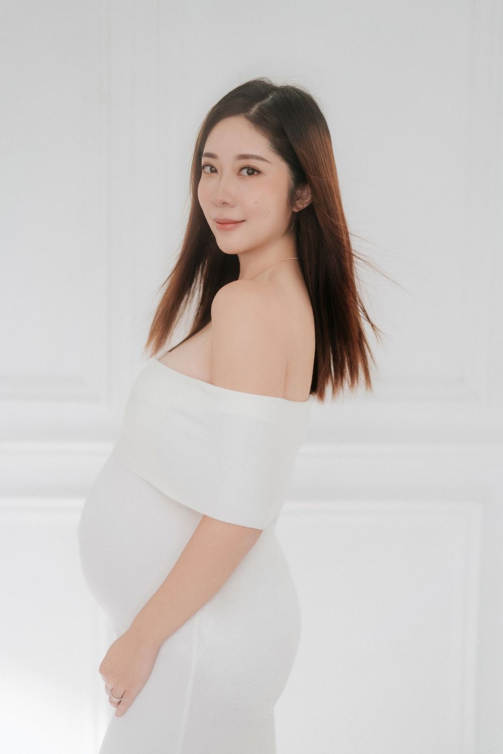 A person in a white off-shoulder top poses gracefully against a light background in a minimalist studio setting.在極簡主義風格的攝影棚內，一位身穿白色露肩上衣的女子在淺