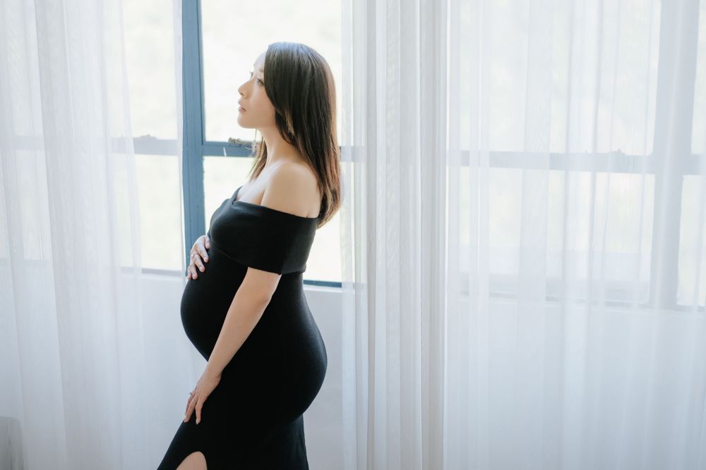 Silhouetted pregnant woman in black dress stands by window with sheer white curtains in soft natural light.在柔和的自然光下，一位身穿黑色連身裙的孕婦站在掛著白色薄紗窗簾的窗
