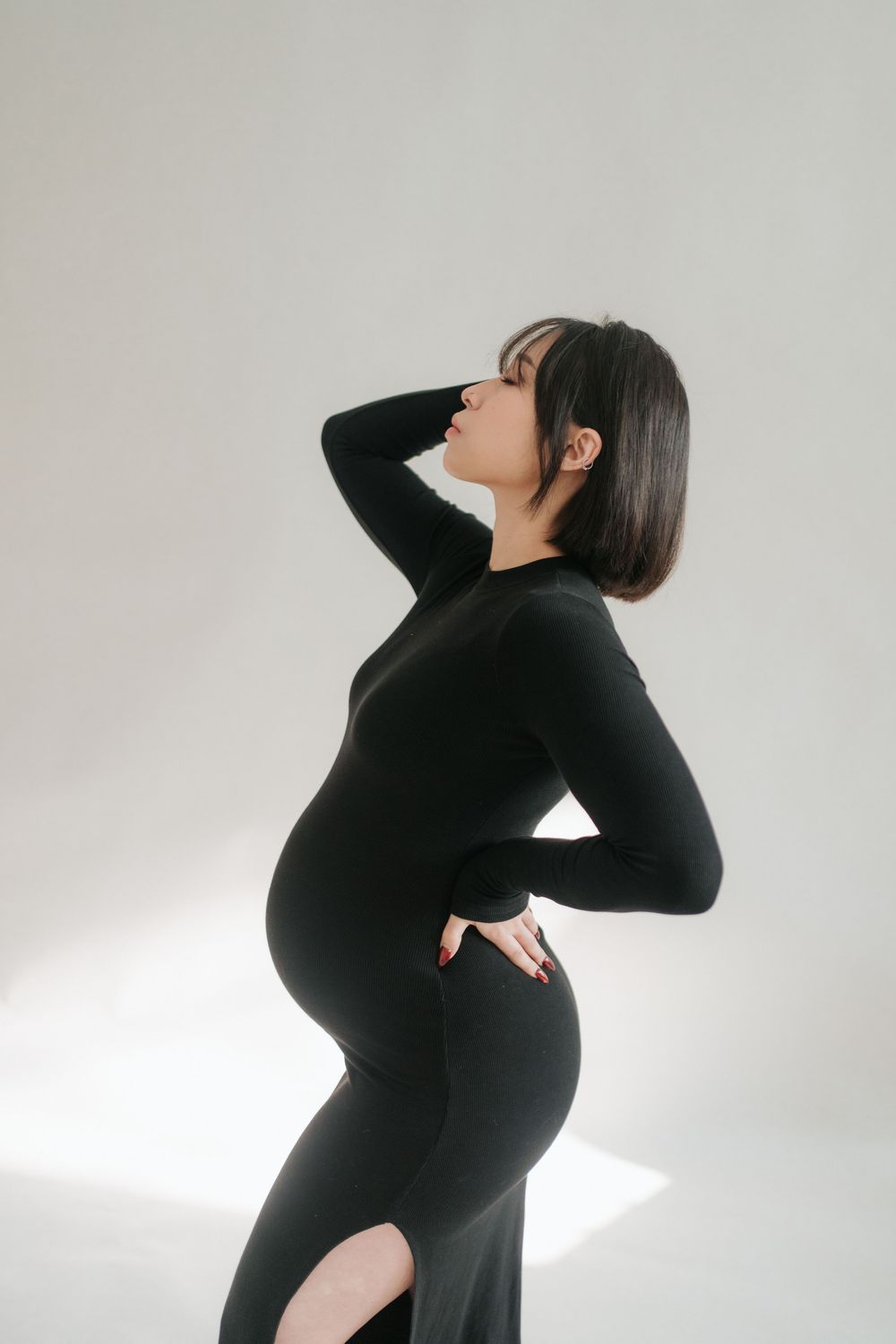 Pregnant woman in black long-sleeve dress poses sideways, hand on back, showcasing baby bump against white background.一位身穿黑色長袖連身裙的孕婦側身擺姿勢
