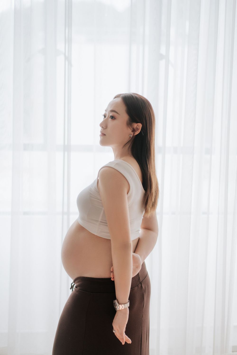 Woman in white top and dark pants poses gracefully against sheer white curtains in a bright interior setting.一位身穿白色上衣和深色褲子的女子在明亮的室內環境中